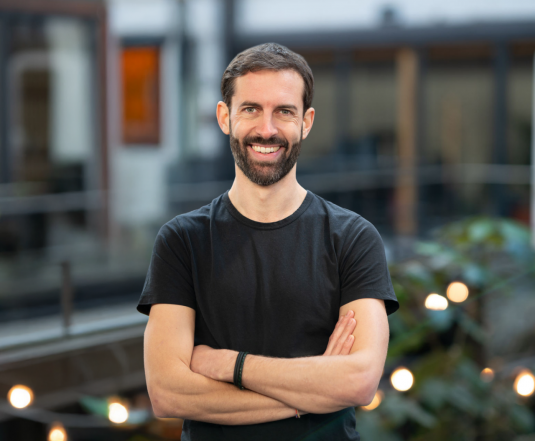 Barnabas Ohst steht mit verschränkten Armen in der Mitte des bildes und lächelt in die Kamera